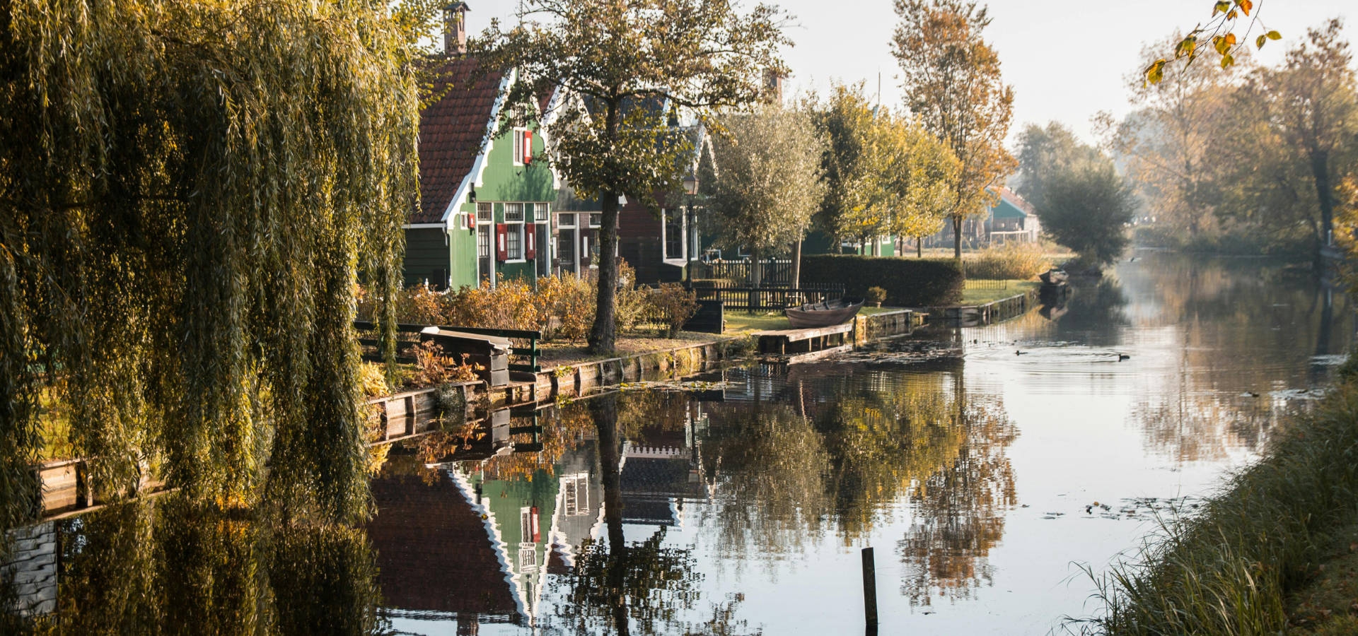 Rustig dorp in herfstkleuren met oude huizen, stil water en bomen in zacht ochtendlicht.