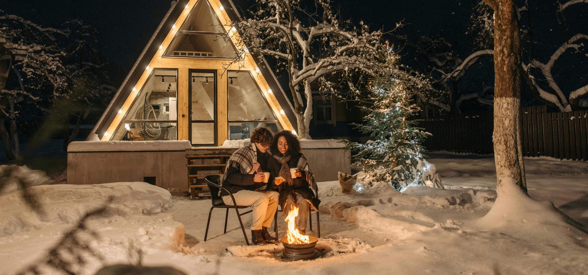 Koppel drinkt warme chocolademelk bij een vuurkorf voor een sfeervol natuurhuisje in de sneeuw.
